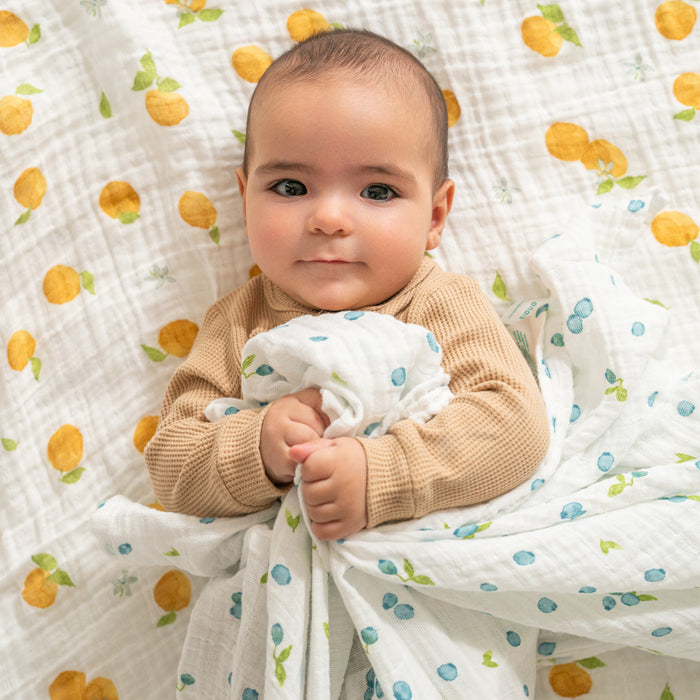 A baby with short dark hair lies on a patterned blanket with yellow and green motifs, dressed in a beige outfit. They are wrapped in an aden + anais organic cotton muslin swaddle from the 4-pack, holding another white blanket with blue and green patterns, gazing upwards gently.