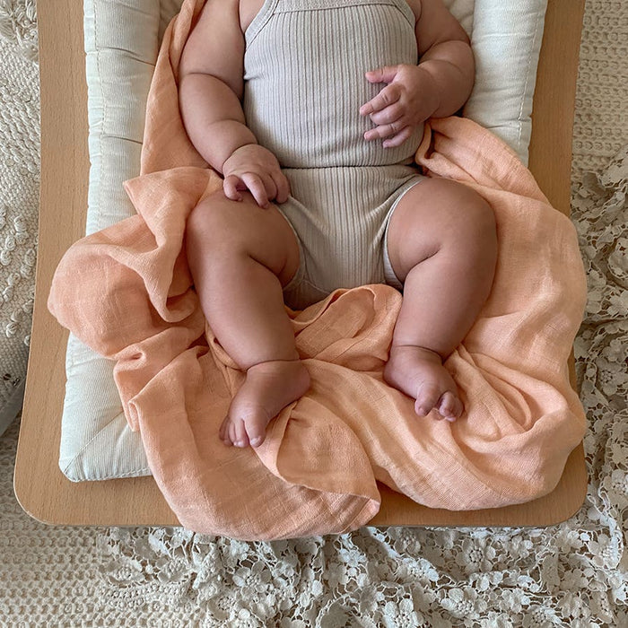A baby in a light beige outfit lies on a cushioned changing pad with an aden + anais organic cotton muslin swaddle blanket, enhancing comfort with its soft texture. The scene is cozy as the baby rests their hands and feet gently on this sustainable swaddle.