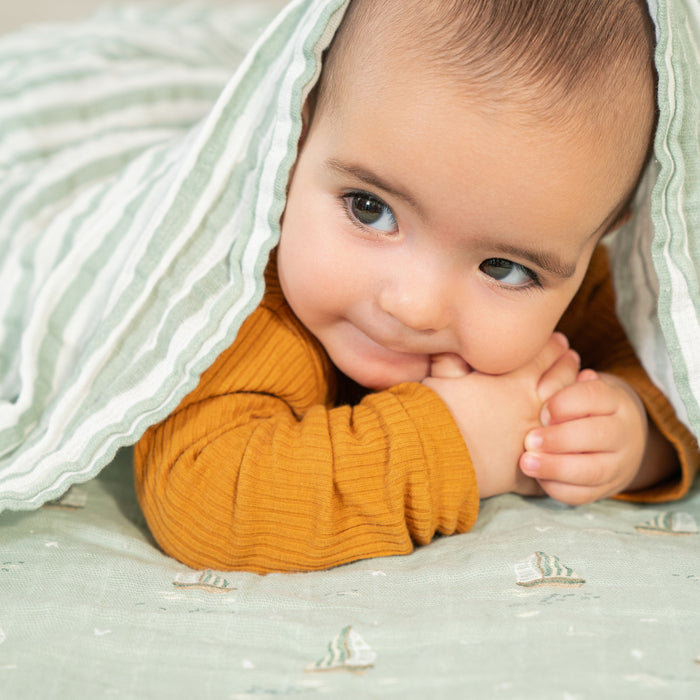 A baby smiles and rests on a green patterned blanket, partially covered with an aden + anais organic cotton muslin swaddle blanket from the 4 pack, dressed in mustard yellow.
