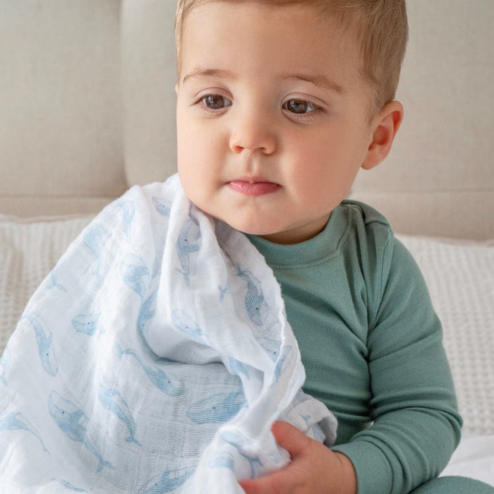 A toddler in a green outfit sits on a soft surface, holding an aden + anais organic cotton muslin swaddle blanket from their 4 pack, featuring a blue whale pattern. Made with GOTS certified materials, the blanket ensures comfort as the child gazes calmly to the side.