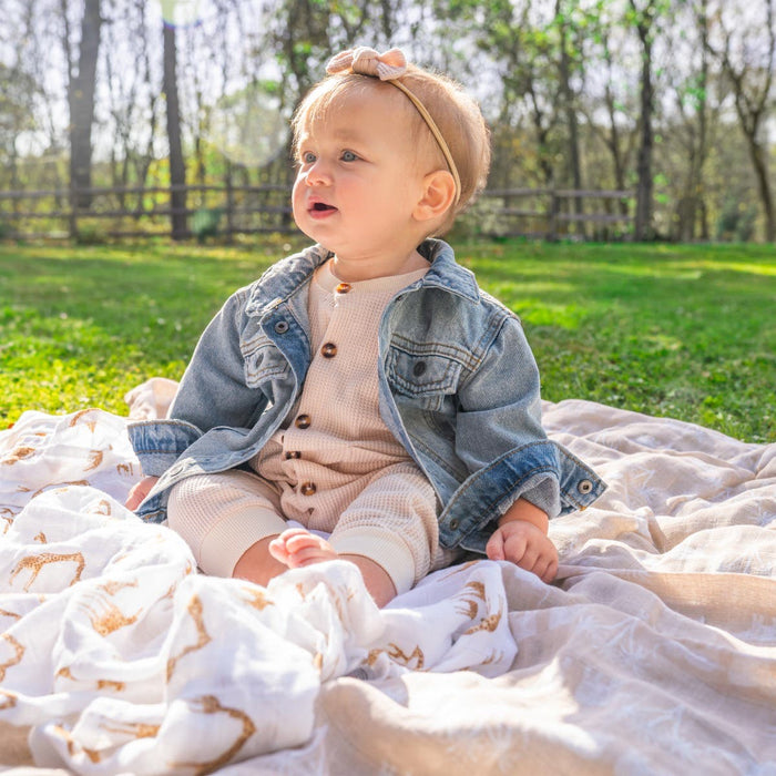 A baby, dressed in a denim jacket and beige outfit, sits on a blanket in nature with clear skies. Wrapped snugly in an aden + anais organic cotton muslin swaddle from their 4-pack, a headband with a bow adds charm to this serene outdoor scene.