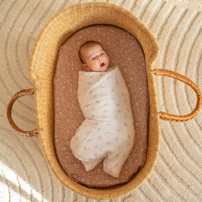 A baby wrapped in a blanket from the aden + anais organic cotton muslin swaddle blanket 4 pack lies in a woven basket with two brown leather handles on a textured carpet, wearing a curious expression.