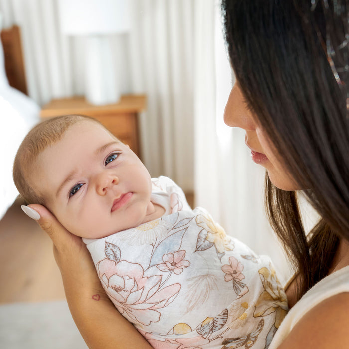 In a softly lit room adorned with white curtains and wooden furniture, a woman with long dark hair gently cradles a content baby wrapped in an aden + anais organic cotton muslin swaddle blanket. The babys light eyes add to the serene backdrop of this tender moment.