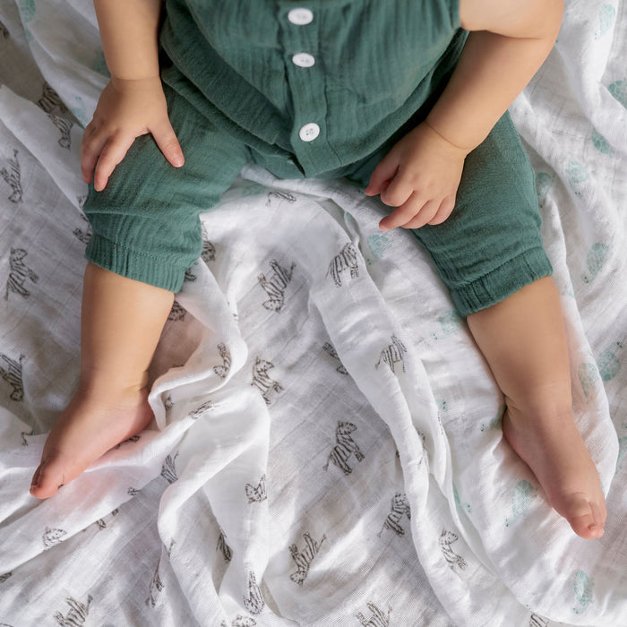 A baby is nestled on an aden + anais zebra-patterned, organic cotton muslin swaddle blanket from a 4-pack. The child, wearing a green buttoned outfit revealing legs and part of the torso, is gently wrapped in the crumpled GOTS-certified blanket.