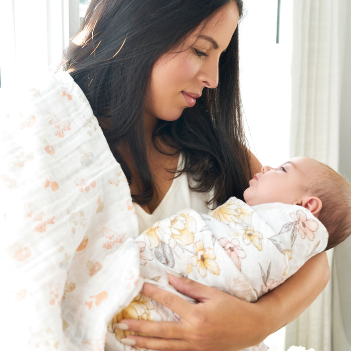 A woman with long dark hair lovingly holds a baby wrapped in an aden + anais organic cotton muslin swaddle blanket featuring florals. Theyre in a bright, sunlit room.