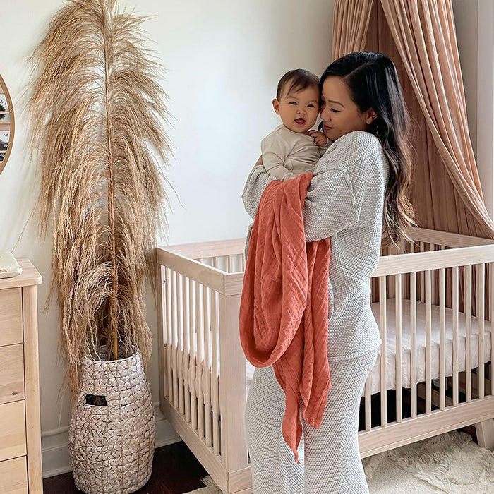 A woman in light-colored pajamas joyfully holds a baby wrapped in an aden + anais organic cotton muslin swaddle blanket. They stand in a cozy nursery with a wooden crib, beige curtains, and a tall vase of pampas grass.