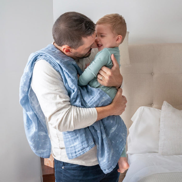 A man warmly holds a smiling child dressed in green, nestled in an aden + anais organic cotton muslin swaddle blanket. They stand near a beige bed with white pillows.