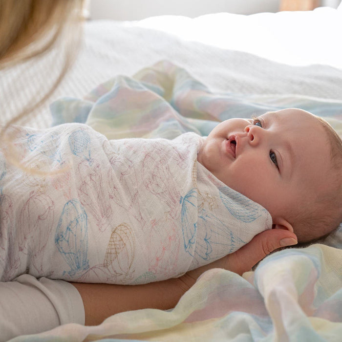 A baby, wrapped in an aden + anais organic cotton muslin swaddle from a 4-pack, lies on pastel bedding, gazing upward. A blonde individual gently cradles the baby.