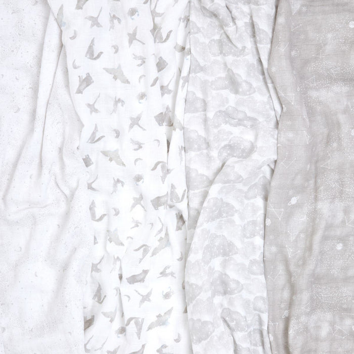 The aden + anais organic cotton muslin swaddle blanket 4 pack includes four GOTS certified blankets. From left to right: white with speckles, white with starfish, white with cloud motifs, and beige with a faint design. All offer a soft, muted look as sustainable baby products.