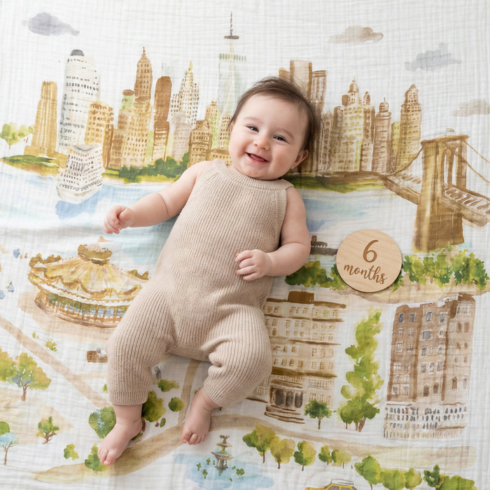 A smiling baby in a beige outfit rests on the aden + anais organic cotton muslin dream blanket, featuring a cityscape print. A round wooden sign next to the baby displays "6 months.