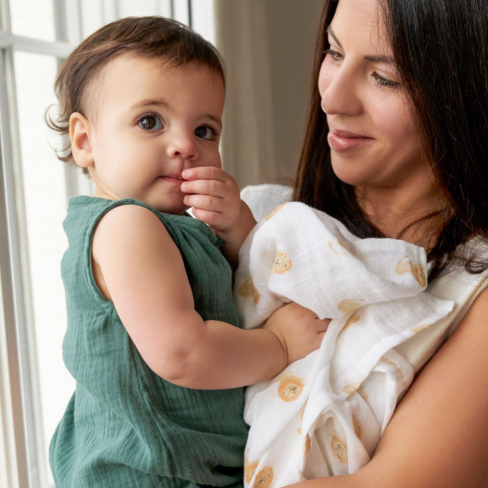 A woman cradles a baby by a window as the child, in a green outfit, touches their lips while gazing at the camera. She smiles warmly, with an aden + anais organic cotton muslin swaddle blanket draped over her shoulder. Natural light fills the room.