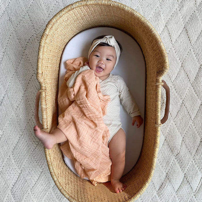 A baby in a headband and light outfit lies in a basket, cozily wrapped in aden + anais soft peach-toned organic cotton muslin swaddle blanket. The little one smiles with their tongue out on a textured, light-hued surface—proof that joy is GOTS certified.