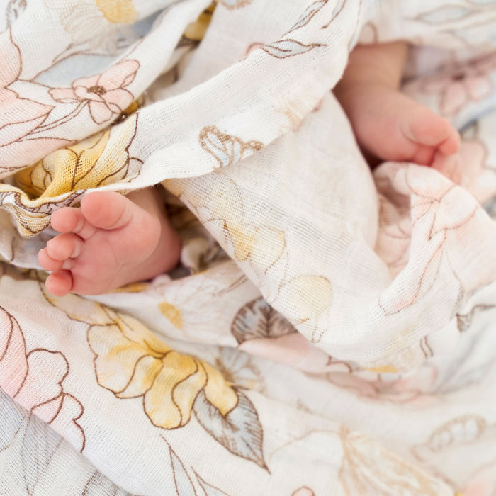 Tiny baby feet peek from a floral-patterned, soft fabric, crafted by aden + anais. This cozy scene is painted with pastel pink, yellow, and green on a 100% organic cotton muslin swaddle blanket 4-pack, providing gentle comfort for delicate skin.