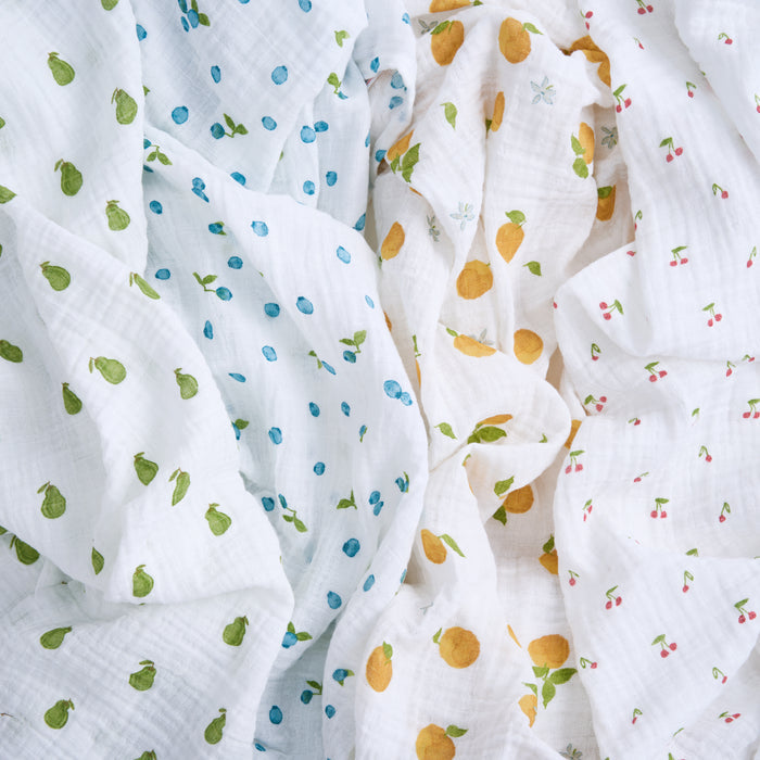 Close-up of crumpled white aden + anais organic cotton muslin swaddle blankets with fruit patterns: green pears on the left, blue blueberries with yellow lemons in the middle, and red cherries on the right. This playful design is perfect for sustainable baby products.