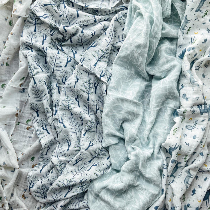 Aden + Anais organic cotton muslin swaddles 4 pack showcases crumpled fabrics with tree branches, leaves, and small animals in blue, green, and white patterns. These GOTS certified blankets overlap for a textured look.