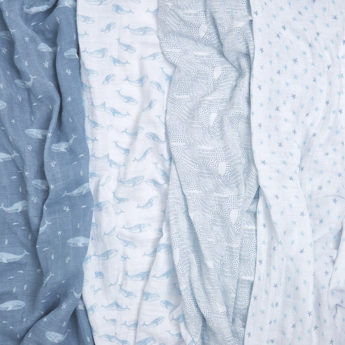 The aden + anais organic cotton muslin swaddle blanket 4 pack features four soft fabrics with unique patterns: blue whales, pale whales, white with gray lines, and white with faint star designs. These GOTS certified blankets showcase a palette from dark blue to light gray and white.