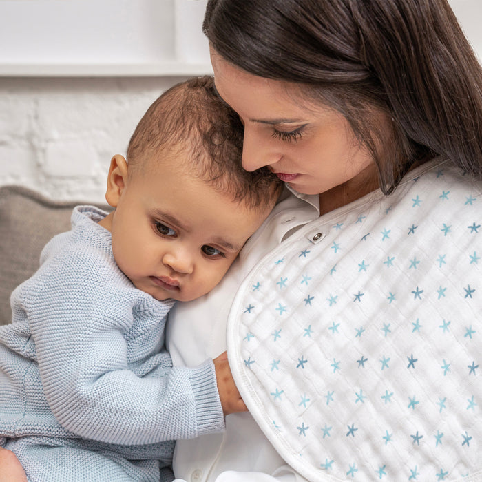 A woman comforts her baby, who’s in a light blue sweater. Draped over her shoulder is an aden + anais organic cotton snap bib with blue stars, part of their 3-pack set. Both share a calm, affectionate moment.