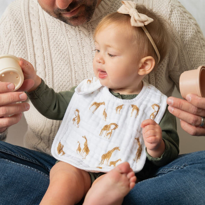 A baby wearing an aden + anais organic cotton snap bib (3 pack) with giraffe designs sits on an adults knee, holding a toy cup. The adult, in a knitted sweater, assists with the toy, creating a warm and cozy scene.