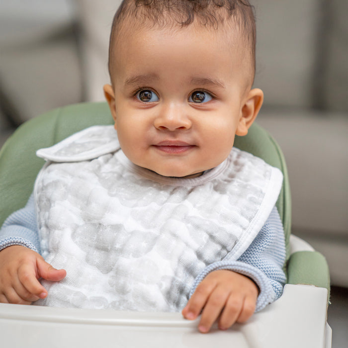 A baby with curly hair and brown eyes sits in a high chair, smiling and wearing an aden + anais organic cotton snap bib from the 3 pack set, along with a light blue sweater.