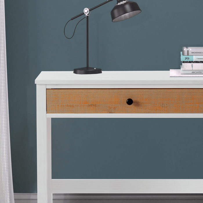 The Child Craft Rockport Desk in Sandstone features a single wooden drawer, paired with a black desk lamp and a small stack of books, all set against a blue wall with white curtains partially visible on the left.