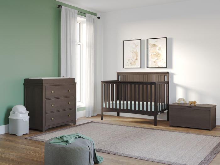 A modern nursery with green and white walls, a Child Craft Scout 4-in-1 Convertible Crib in Dapper Gray, matching dresser and toy chest, diaper pail, framed leaf art, pouf, and a large beige rug.