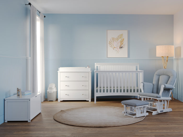 A bright nursery with light blue walls features the Child Craft Scout 4-in-1 Convertible Crib in Matte White, a dresser, rocking chair with ottoman, storage chest, round beige rug, floor lamp, and framed leaf artwork. Sunlight streams through the window.