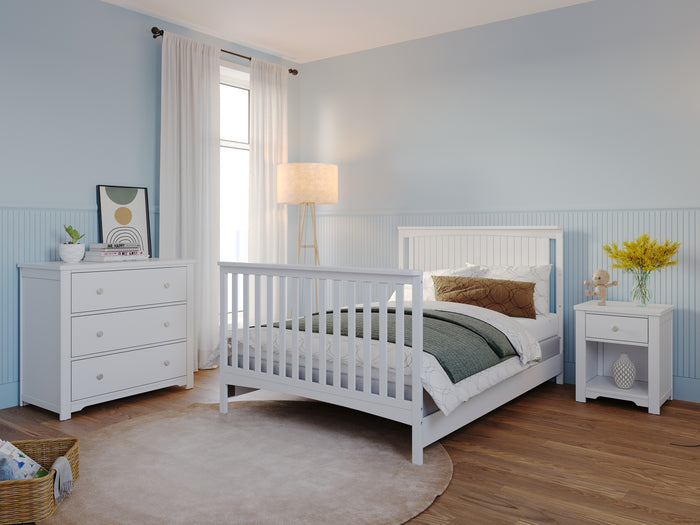 A cozy bedroom features a Child Craft Scout 4-in-1 Convertible Crib in Matte White, a matching white bed, nightstand with yellow flowers, dresser, floor lamp, sheer-curtained window, round beige rug, all set against light blue walls.