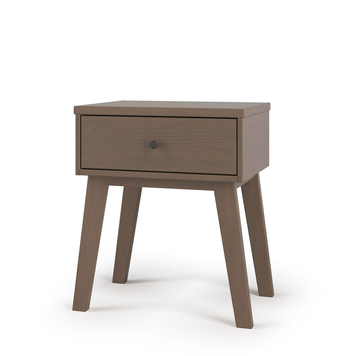 The Child Craft Soho Nightstand in Dusty Heather is a mid-century modern nursery piece featuring one drawer with a round black knob, four angled legs, and a smooth light brown finish, shown against a plain white background.