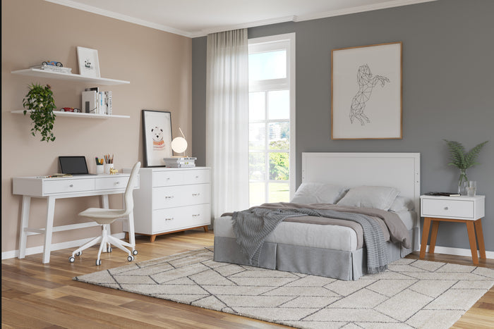 A modern bedroom features a gray accent wall, neatly made bed, white desk and chair, dresser, wall art, and the Child Craft Soho Nightstand in White/Natural. Sunlight from a large window with sheer curtains brightens the cozy, organized space.