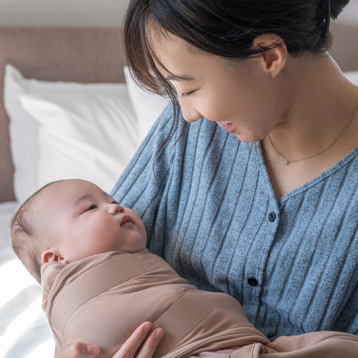 A woman in a blue sweater smiles while holding a baby wrapped in the HALO SuperSoft Bamboo Swaddle TOG 2.0. They sit indoors on a bed with white pillows in the background.
