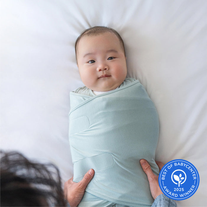 A baby lies on a white bed in a light blue HALO Bedside Sleeper Bassinet Bundle, looking up as an adult gently holds them. A blue Best of BabyCenter 2025 Award Winner badge appears in the lower right corner.