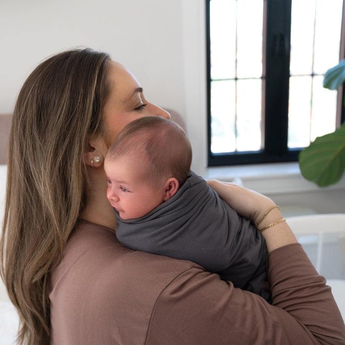 A woman with long brown hair holds a newborn wrapped in a gray HALO Swaddle SuperSoft Bamboo TOG 2.0, close to her shoulder, in a softly lit room with a window and green plant in the background.