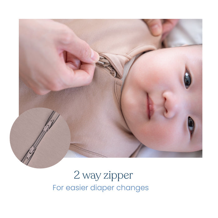 A close-up of an adult hand zipping up the beige HALO SuperSoft Bamboo Swaddle TOG 2.0, made from soft bamboo fabric with a two-way zipper for easier diaper changes. Inset shows a detailed view of the zipper: 2-way zipper for convenience.