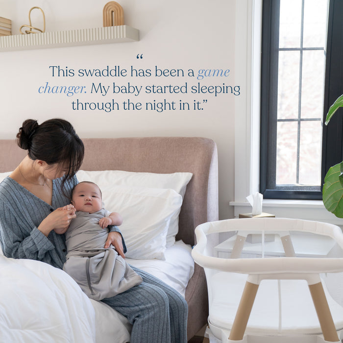 A woman sits on a bed holding a baby wrapped in a HALO Swaddle SuperSoft Bamboo TOG 2.0. A bassinet is nearby. On the wall, text reads: “This swaddle has been a game changer. My baby started sleeping through the night in it.”.