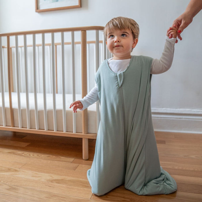 A toddler in a sage green HALO SuperSoft Bamboo Wearable Blanket TOG 1.5 kneels on a wooden floor, holding an adult's hand, with a wooden crib featuring white bars against a white wall in the background.