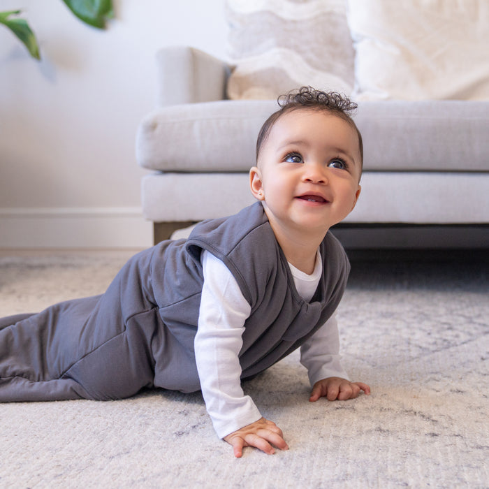 A smiling baby wearing the HALO SuperSoft Bamboo Wearable Blanket TOG 1.5 with a 2-way zipper crawls on a soft carpet in a cozy living room, with a beige sofa in the background.