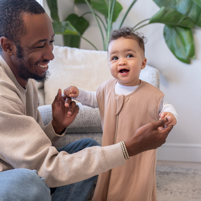 A smiling man helps a happy baby stand in a cozy living room accented by green plants and a light armchair. The baby wears the HALO SuperSoft Bamboo Wearable Blanket TOG 1.5, featuring buttery bamboo fabric and a convenient 2-way zipper.