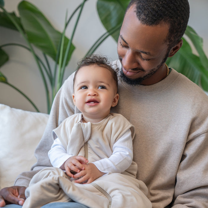 A smiling adult holds a happy baby in front of green leaves indoors; the baby is cozy in the HALO SuperSoft Bamboo Wearable Blanket TOG 1.5.