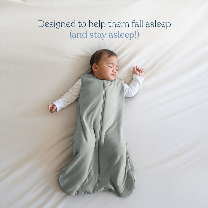 A baby sleeps peacefully on a white bed in the HALO SuperSoft Bamboo Wearable Blanket TOG 1.5, featuring a cozy gray design and 2-way zipper. Text reads: Designed to help them fall asleep (and stay asleep!).