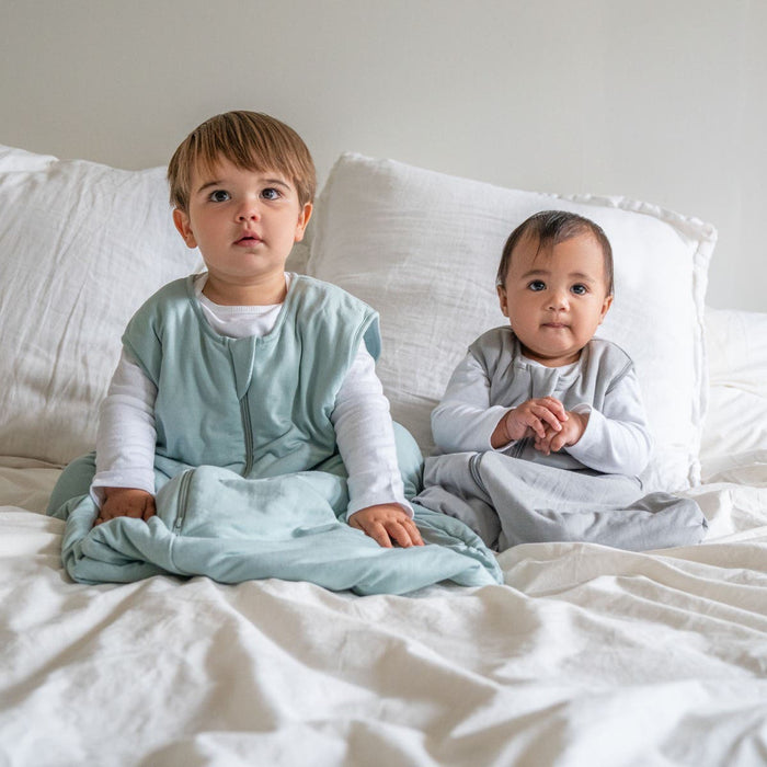 Two young children sit on a bed with white bedding, both wearing HALO SuperSoft Bamboo Wearable Blanket TOG 1.5 sleep sacks and long-sleeve shirts, looking slightly upward.