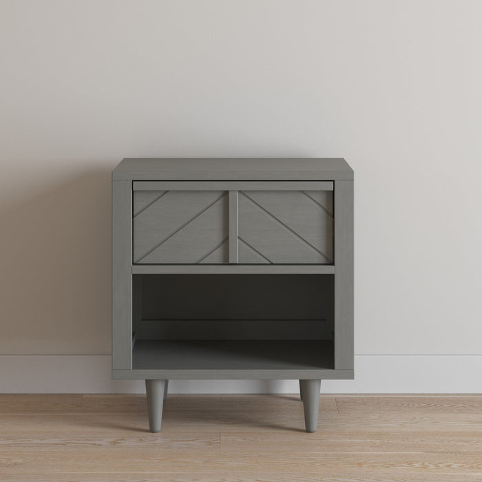 The Child Craft Surrey Hill Nightstand in Lunar Gray features angled legs, a decorative drawer with diagonal lines, and an open shelf below, displayed against a plain white wall on a light wood floor.