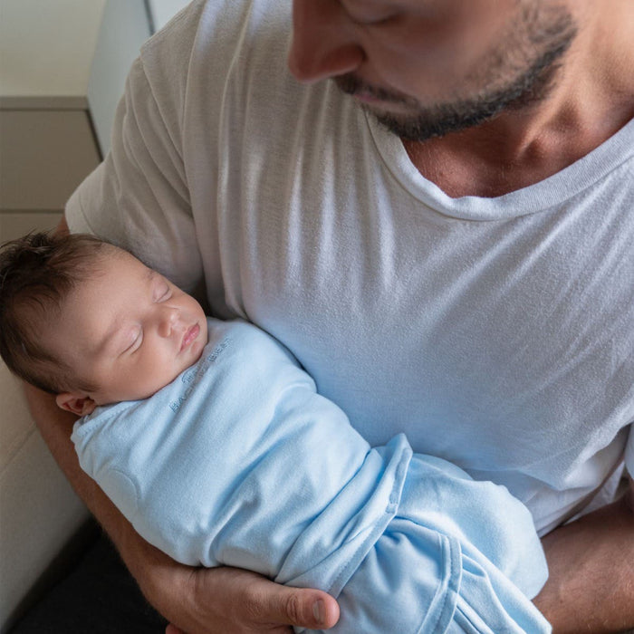 A man in a white shirt holds a sleeping baby wrapped in a HALO Swaddle 100% Cotton TOG 1.5. The baby rests peacefully in his arms, benefiting from the healthy hip development design, as they sit comfortably indoors.