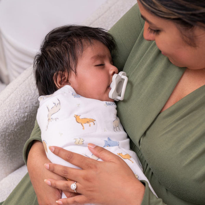 A woman in a green top cradles a sleeping baby swaddled in a HALO Swaddle 100% Cotton TOG 1.5 with animal prints, using the adjustable wings for healthy hip development. The baby rests peacefully with a pacifier, creating a warm and tender scene.