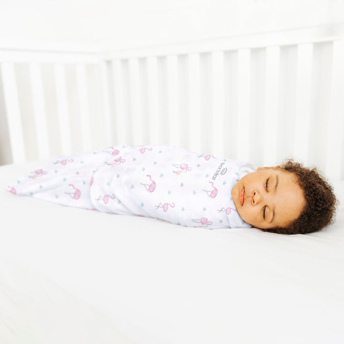 A baby sleeps peacefully in a HALO Swaddle 100% Cotton TOG 1.5 with pink butterfly patterns for healthy hip development. The cribs white bars frame the serene scene, highlighting the babys curly hair on the white sheet.