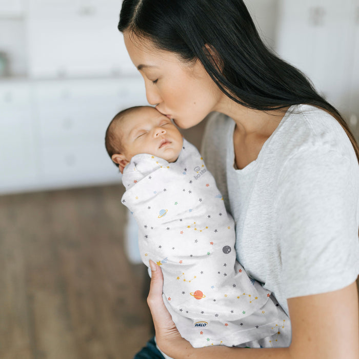 A woman gently kisses a sleeping baby on the forehead, snug in a HALO Swaddle 100% Cotton TOG 1.5 with adjustable wings. The baby is wrapped in a light grey blanket adorned with colorful planets and stars, indoors, against a background of blurred furnishings.