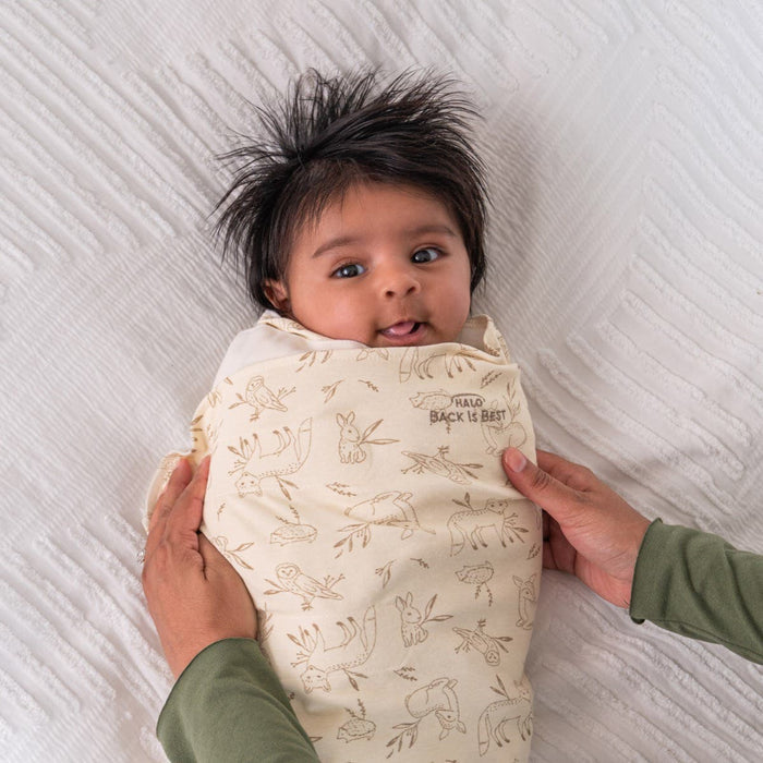 A dark-haired baby is snugly swaddled in a HALO Swaddle 100% Cotton TOG 1.5 with charming animal prints. Resting on a textured white surface, the swaddles adjustable wings ensure comfort and promote healthy hip development as two hands gently cradle the little one.