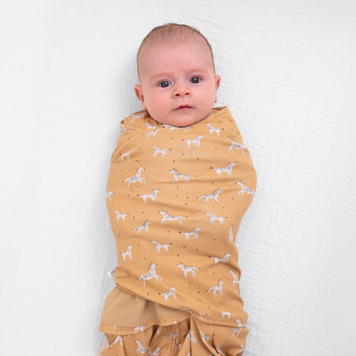 A baby is swaddled in the HALO Fall/Winter Swaddle Bundle’s mustard yellow organic cotton muslin blanket with white zebra prints, lying on a white surface and looking up calmly.