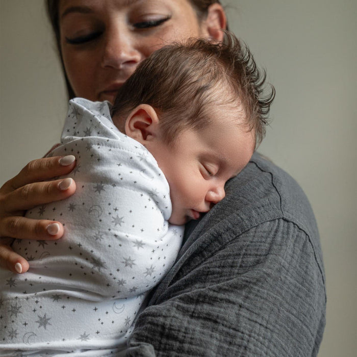 A woman gently cradles a sleeping baby wrapped in a HALO Swaddle 100% Cotton TOG 1.5, adorned with stars and moons. The swaddles adjustable wings ensure comfort as they rest together in the serene settings soft, warm glow.