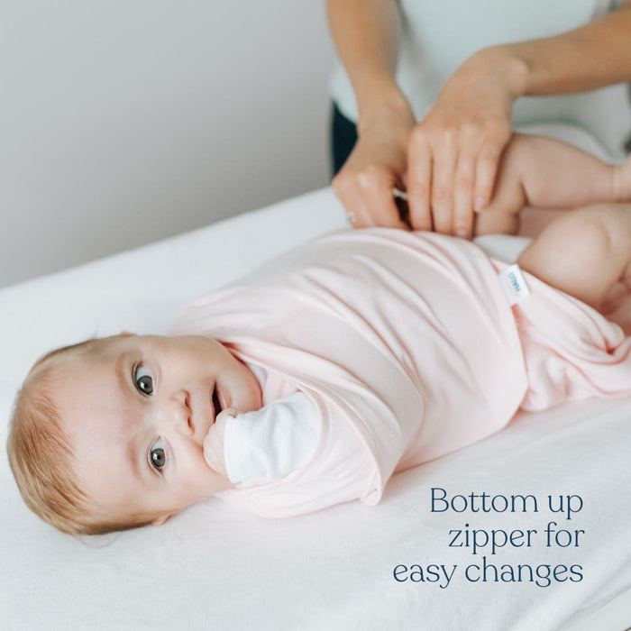 A baby in a light pink HALO Swaddle 100% Cotton TOG 1.5 lies on a changing table while an adult changes their diaper. Text reads: Bottom-up zipper for easy changes and adjustable wings for a snug fit.