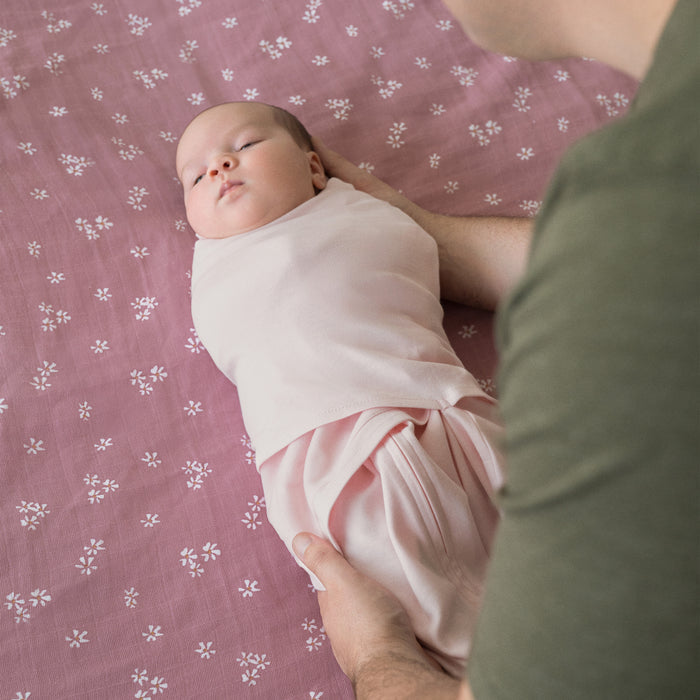 A baby in a light pink HALO Swaddle 100% Organic Cotton TOG 1.5 rests on a mauve sheet with small white floral patterns, gently held by an adult in a green shirt for comfort and security.